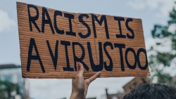 A placard that says "Racism is a virus too", held by a black hand with a tree at the back. Source: Unplash website (CC)